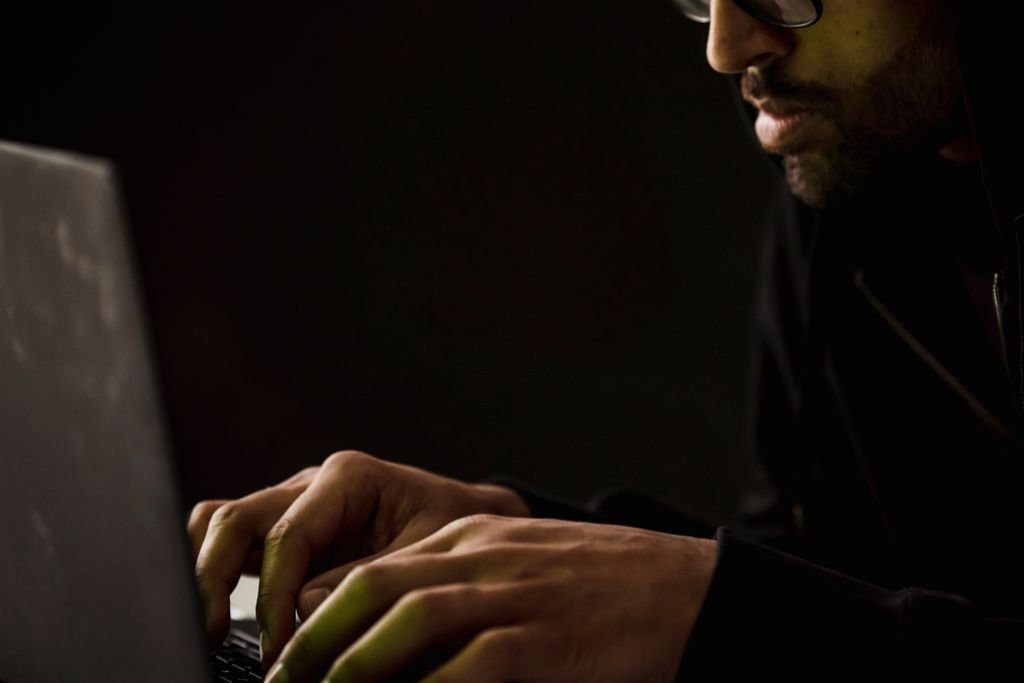 Crop concentrated bearded male in eyeglasses and black clothes browsing modern netbook in dark room